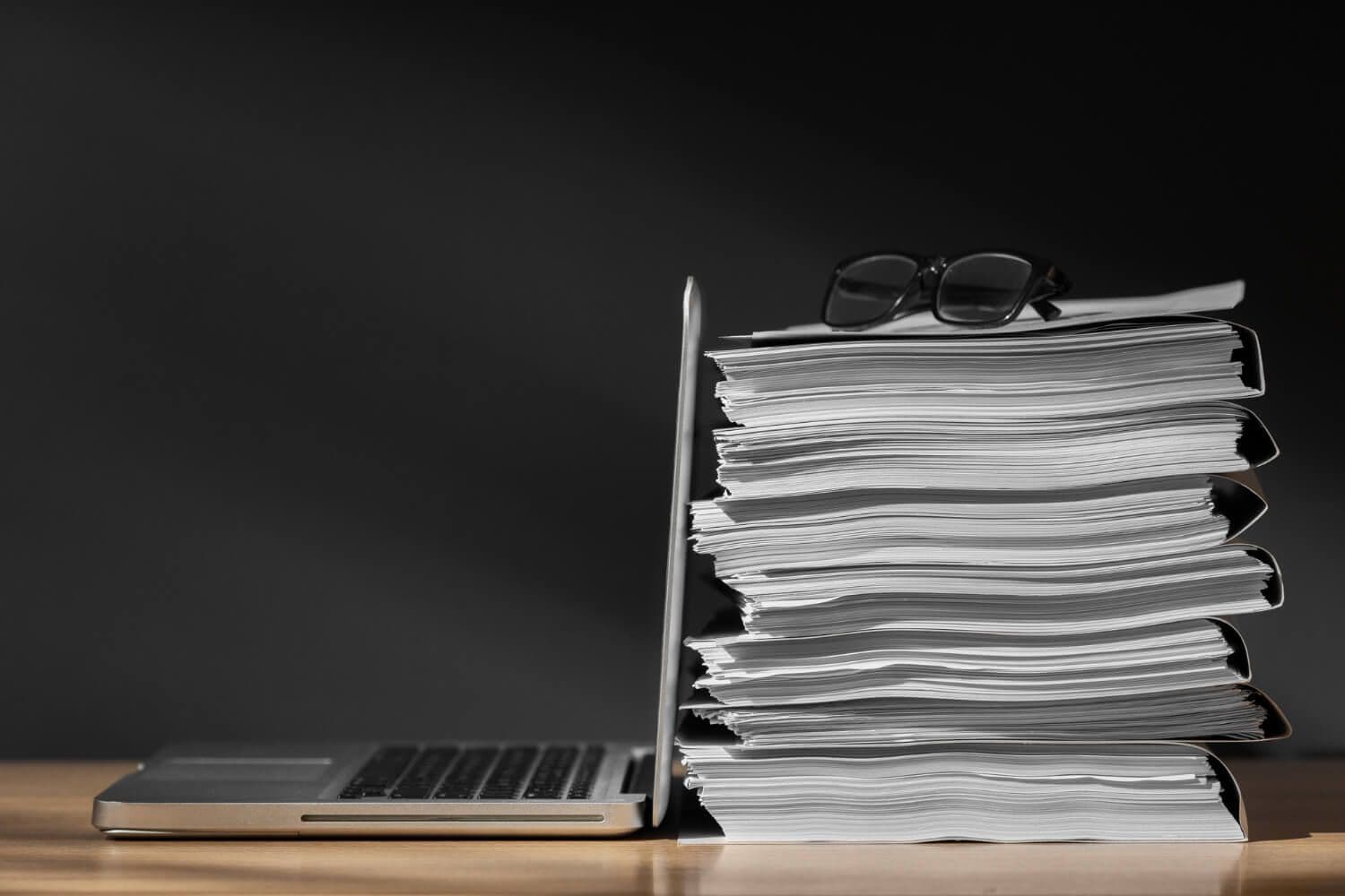 laptop i leżąca za nim kupa dokumentów